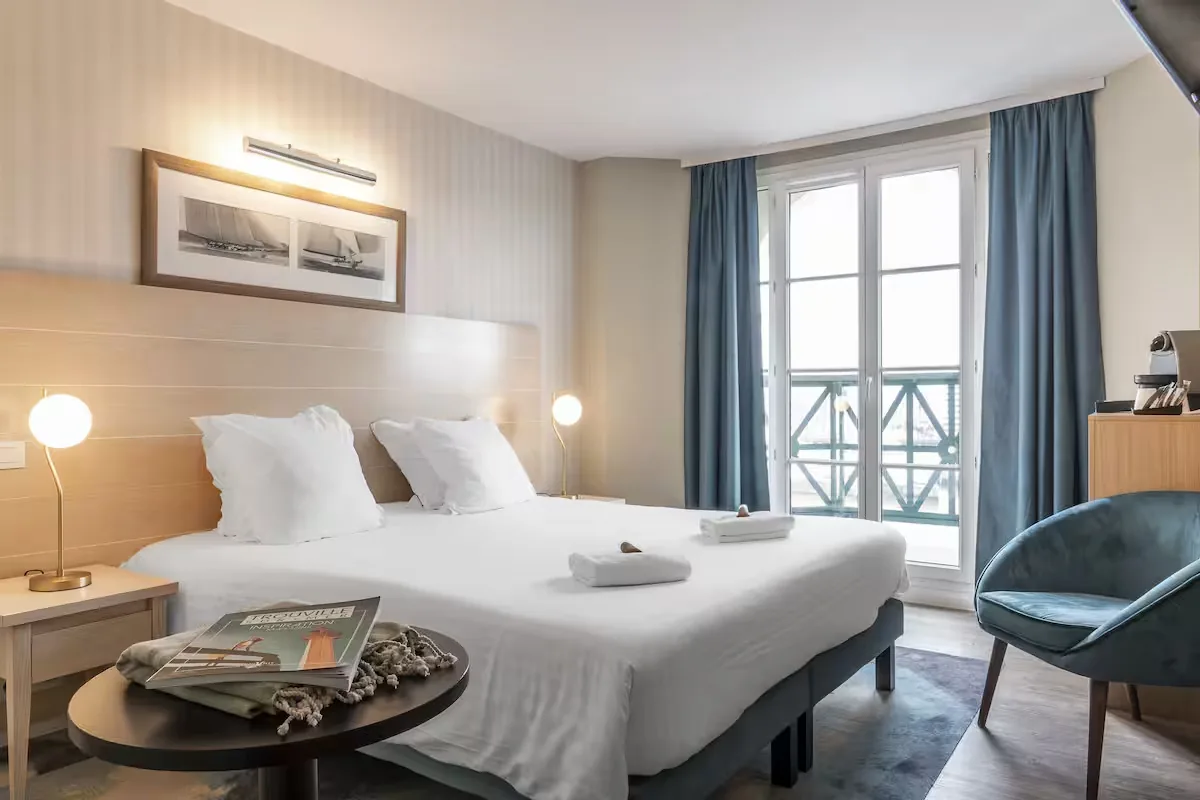 Chambre avec lit double, tête de lit en bois clair, fauteuil bleu canard et porte-fenêtre ouvrant sur un balcon face au port