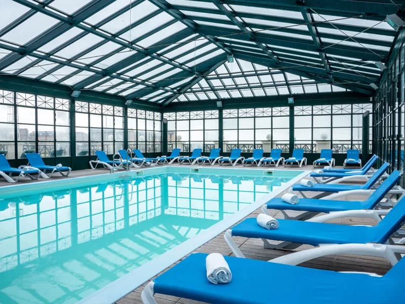 Piscine intérieure sous une verrière à charpente vert bouteille, transats bleus alignés le long du bassin