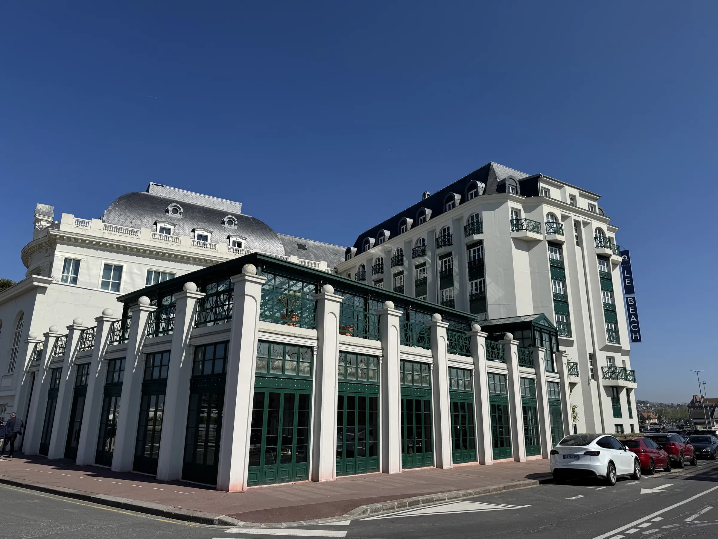 Façade du Beach Hôtel à Trouville-sur-Mer : bâtiment blanc à toitures d’ardoise et balcons vert bouteille, véranda à colonnes en rez-de-chaussée, face au port