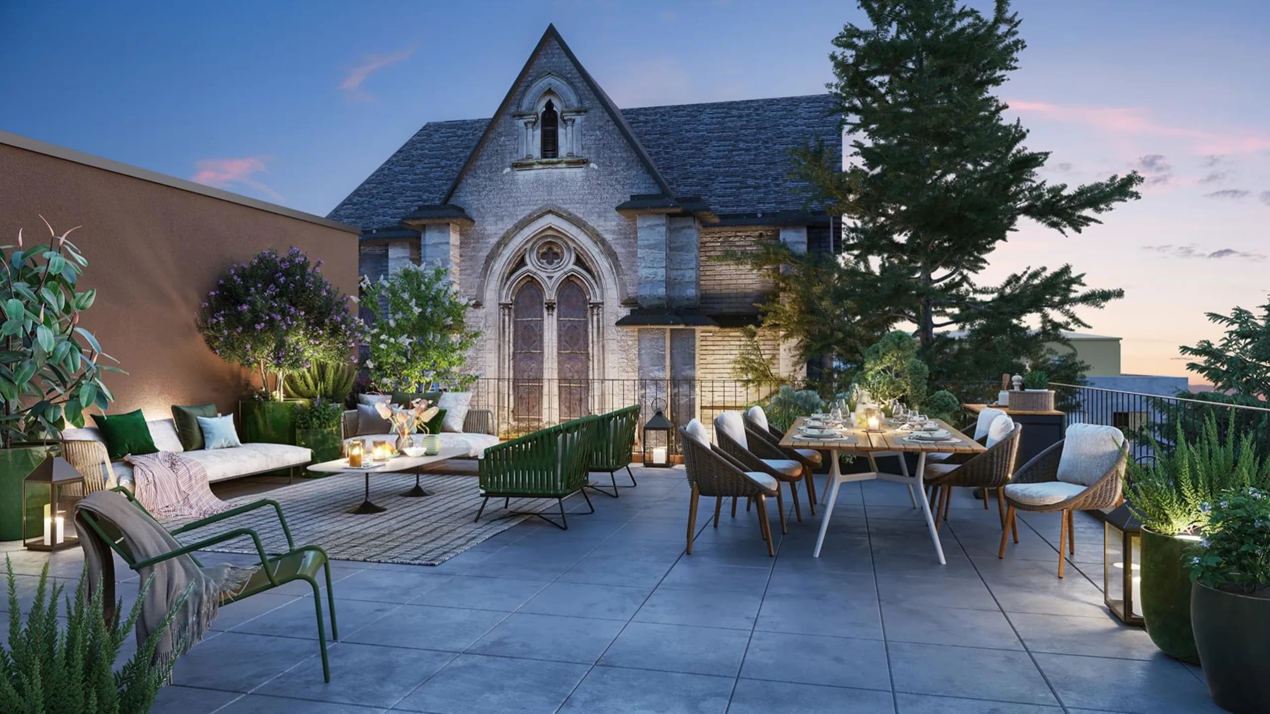 Terrasse d’un appartement au crépuscule, meublée d’un salon d’extérieur et d’une grande table dressée, face à la chapelle en pierre éclairée du domaine