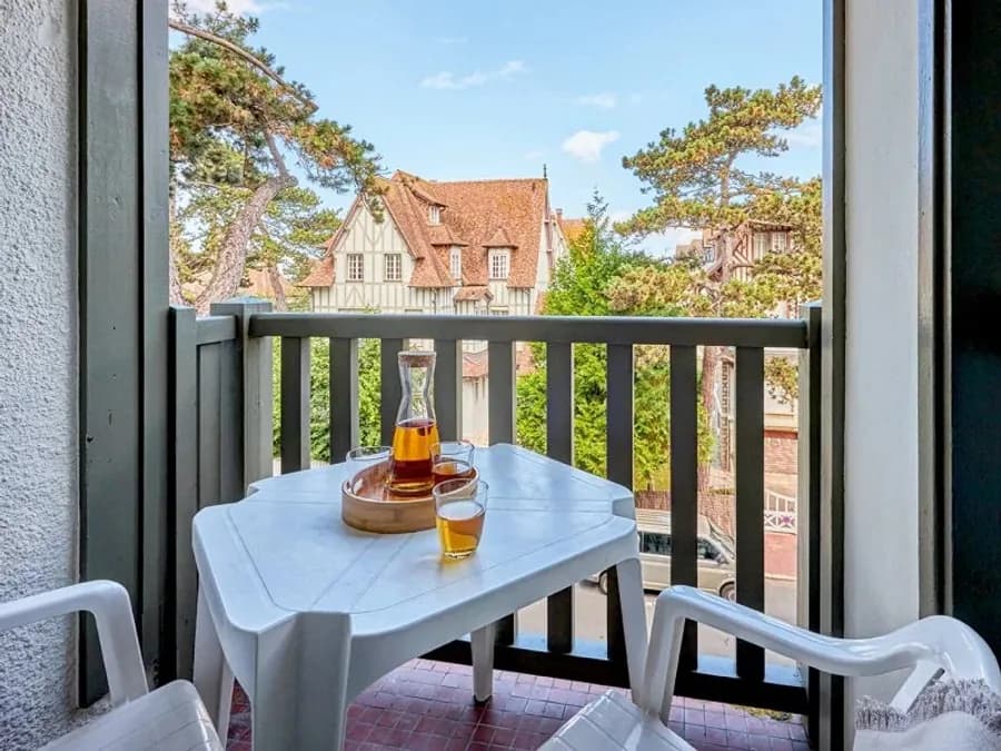 Balcon d’un appartement : table et fauteuils de jardin face à une villa normande à colombages, pins maritimes au-dessus