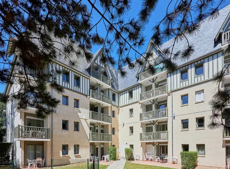Façade des Embruns à Deauville : immeuble à colombages normands et pignons d’ardoise, balcons en bois sur cour arborée, vue sous les pins