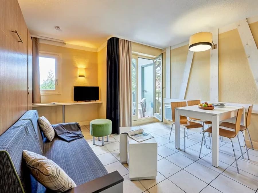 Séjour d’un appartement sous comble : canapé-lit, coin repas, télévision et porte-fenêtre donnant sur le balcon