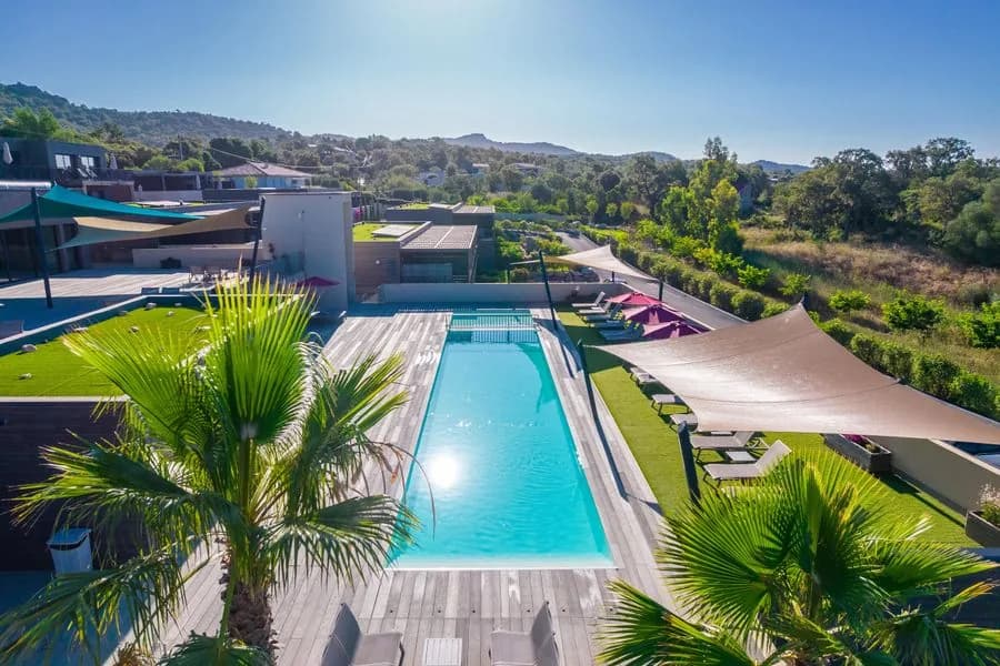 La piscine commune bordée d’une terrasse en bois, voiles d’ombrage et transats, palmiers au premier plan et maquis en arrière-plan