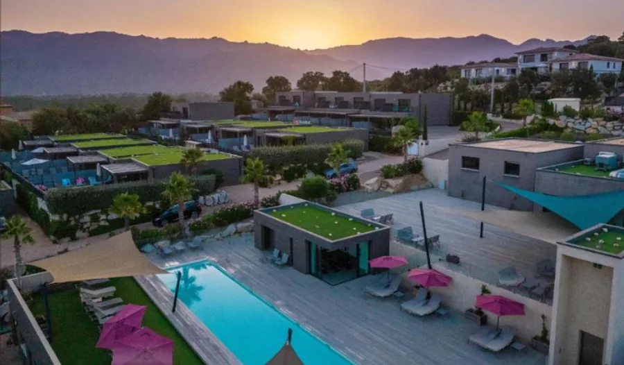 Vue aérienne des Terrasses d’Arsella au coucher du soleil : villas à toitures-terrasses végétalisées échelonnées sur le coteau, piscine et parasols fuchsia au premier plan, montagnes corses à l’horizon