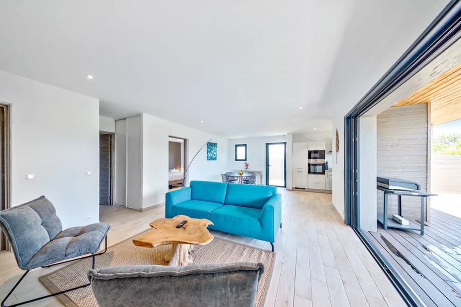 Séjour traversant d’une villa : canapé turquoise, table basse en bois brut, cuisine ouverte au fond et baie coulissante ouverte sur la terrasse