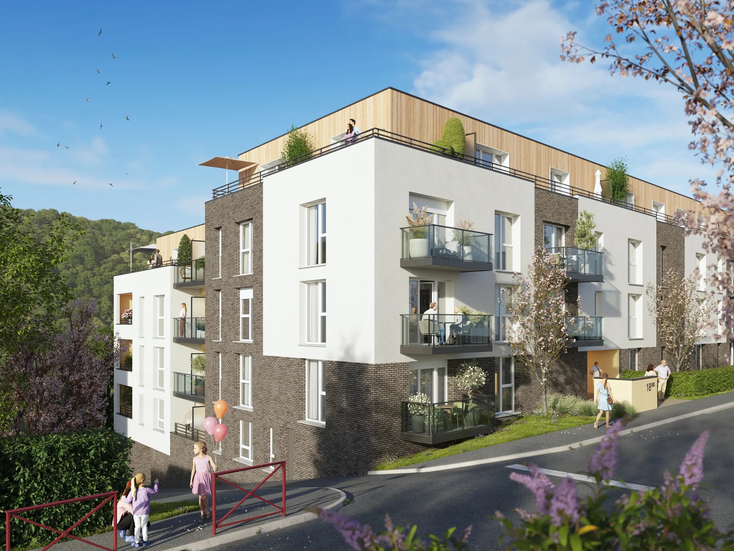 Perspective de Villa Bovary en angle de rue : façade blanche et brique brune, attique bardé de bois, balcons plantés et coteau boisé de la Vallée du Cailly en arrière-plan